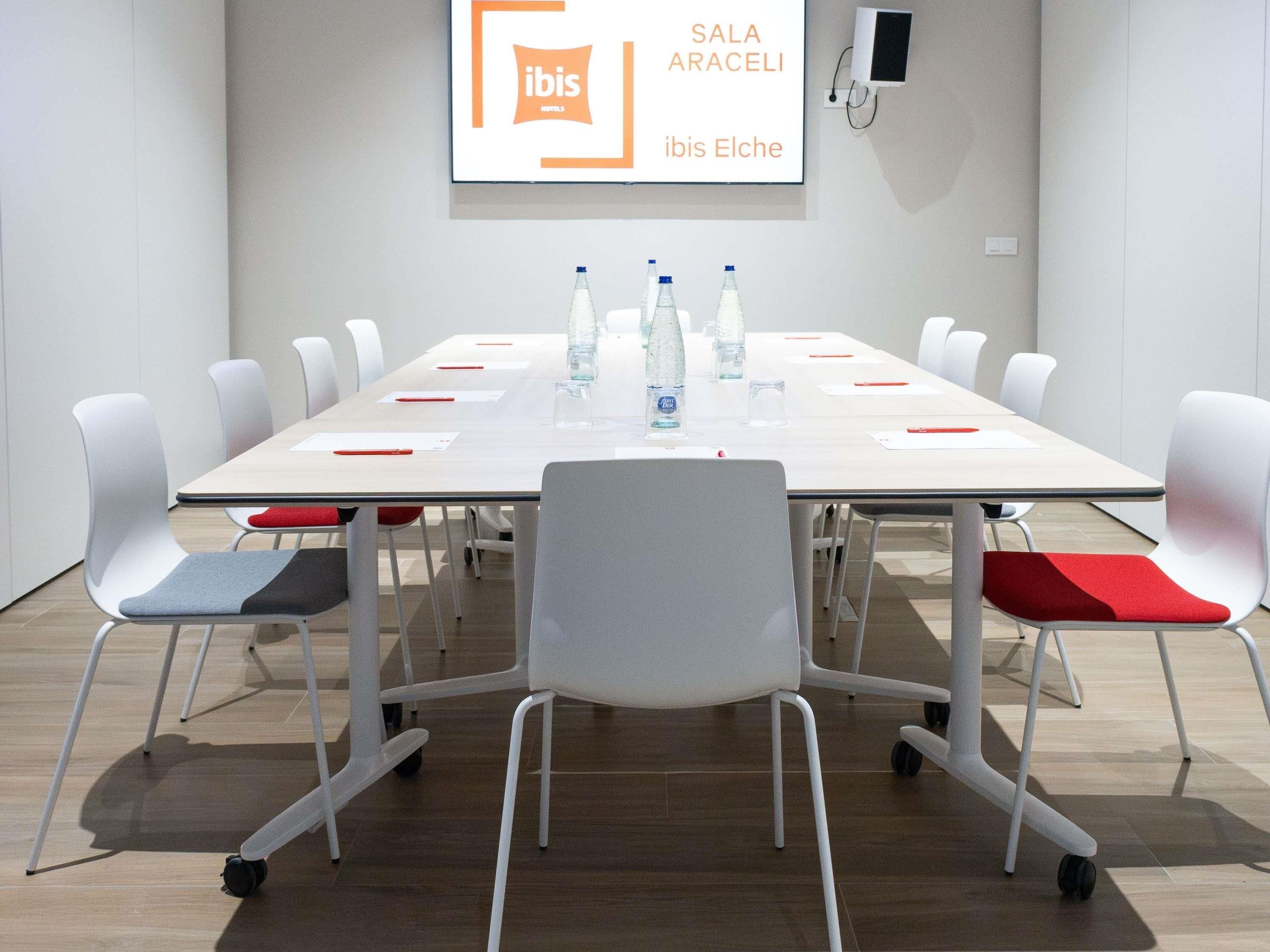 Een vergaderruimte met een witte tafel, rode en grijze stoelen, waterflesjes en een bord met de tekst 'SALA ARACELI ibis Elche'.