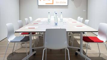 Een vergaderruimte met een witte tafel, rode en grijze stoelen, waterflesjes en een bord met de tekst 'SALA ARACELI ibis Elche'.