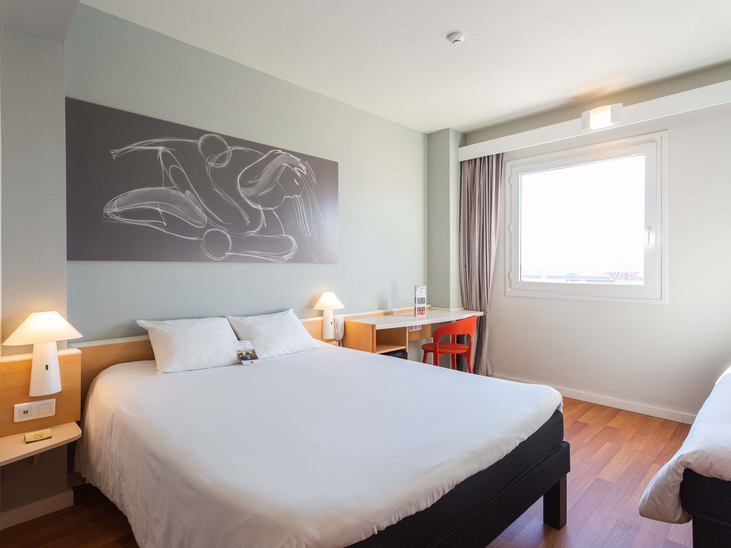 A hotel room with a bed, bedside lamps, a desk, a chair, and a window with curtains.