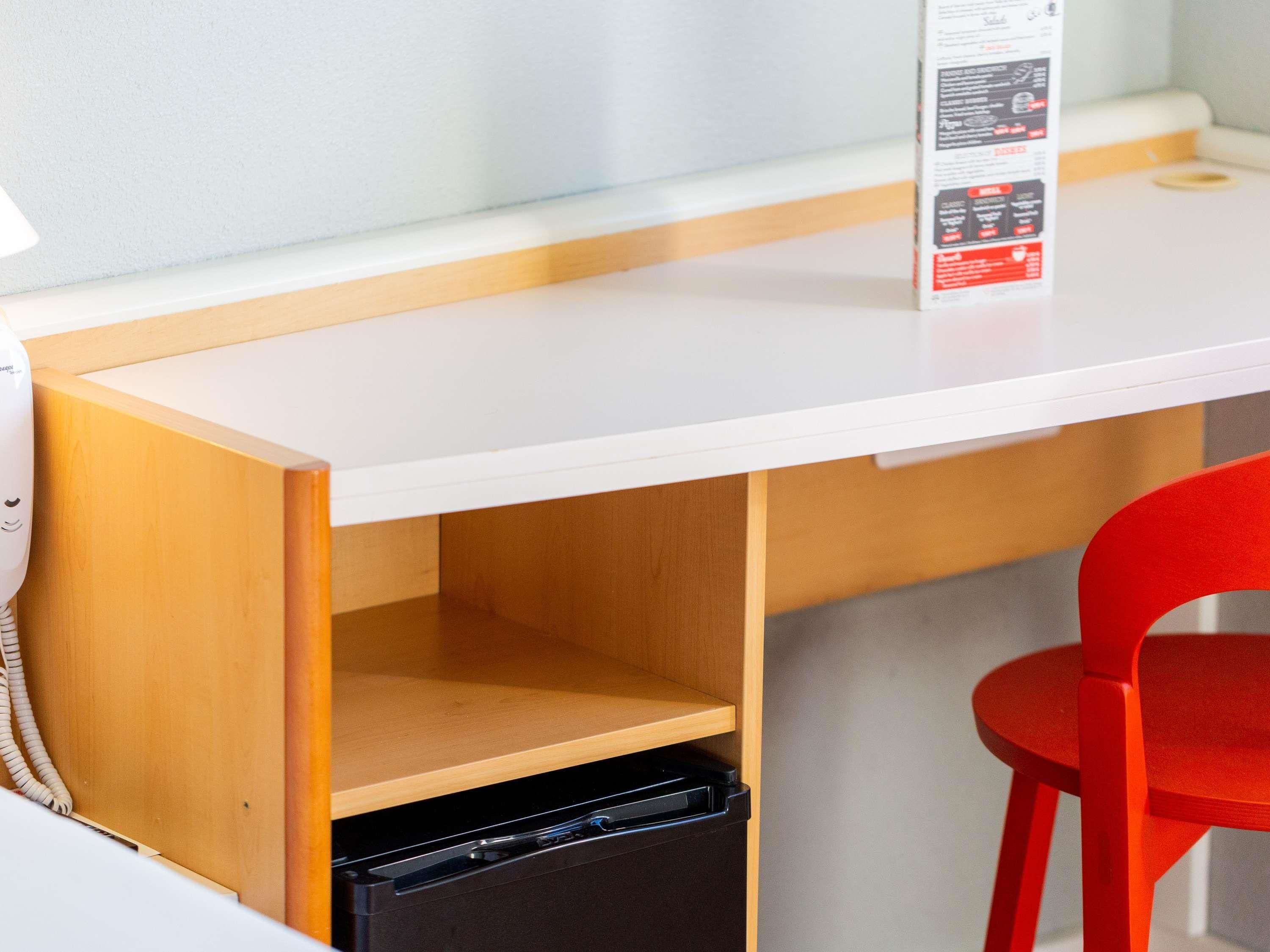 A white desk with a black trash bin underneath, a red chair, and a menu stand on the desk.