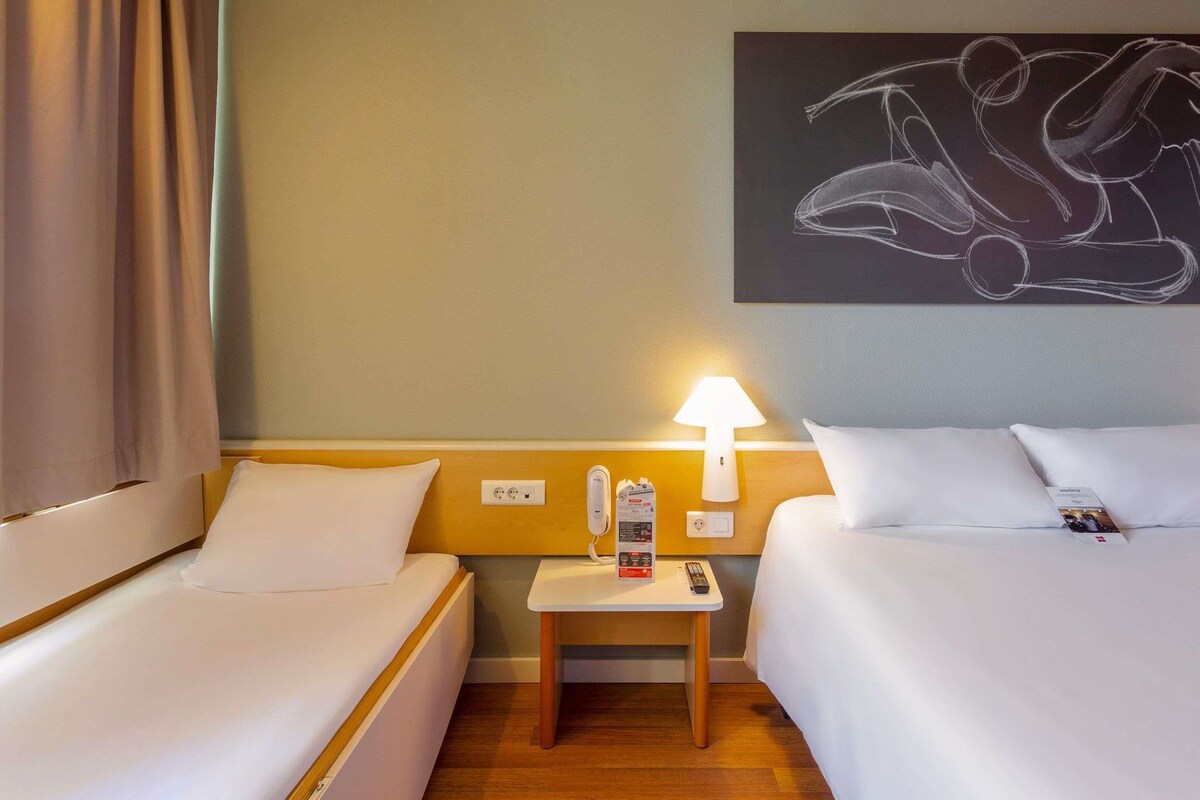 A hotel room with two beds, a nightstand, a lamp, a painting, and a view of the outside.