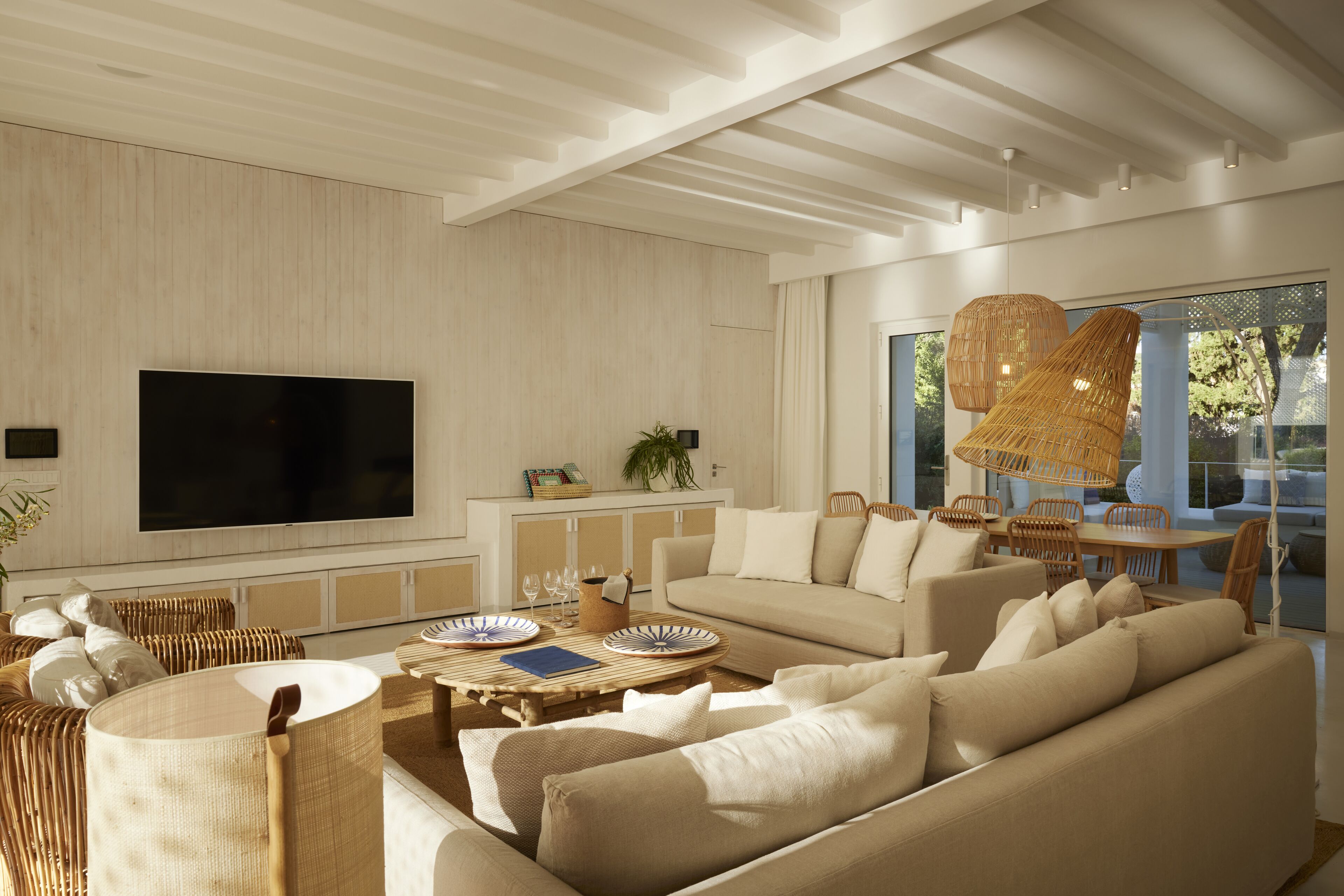 A modern living room with a large flat-screen TV, comfortable sofas, a dining area with wicker furniture, and large windows providing natural light.