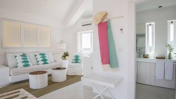 Una sala de estar moderna y bien iluminada con un sofá blanco, dos mesas redondas de centro y una pequeña mesita auxiliar blanca. Se ve una cocina al fondo con armarios blancos y una encimera.