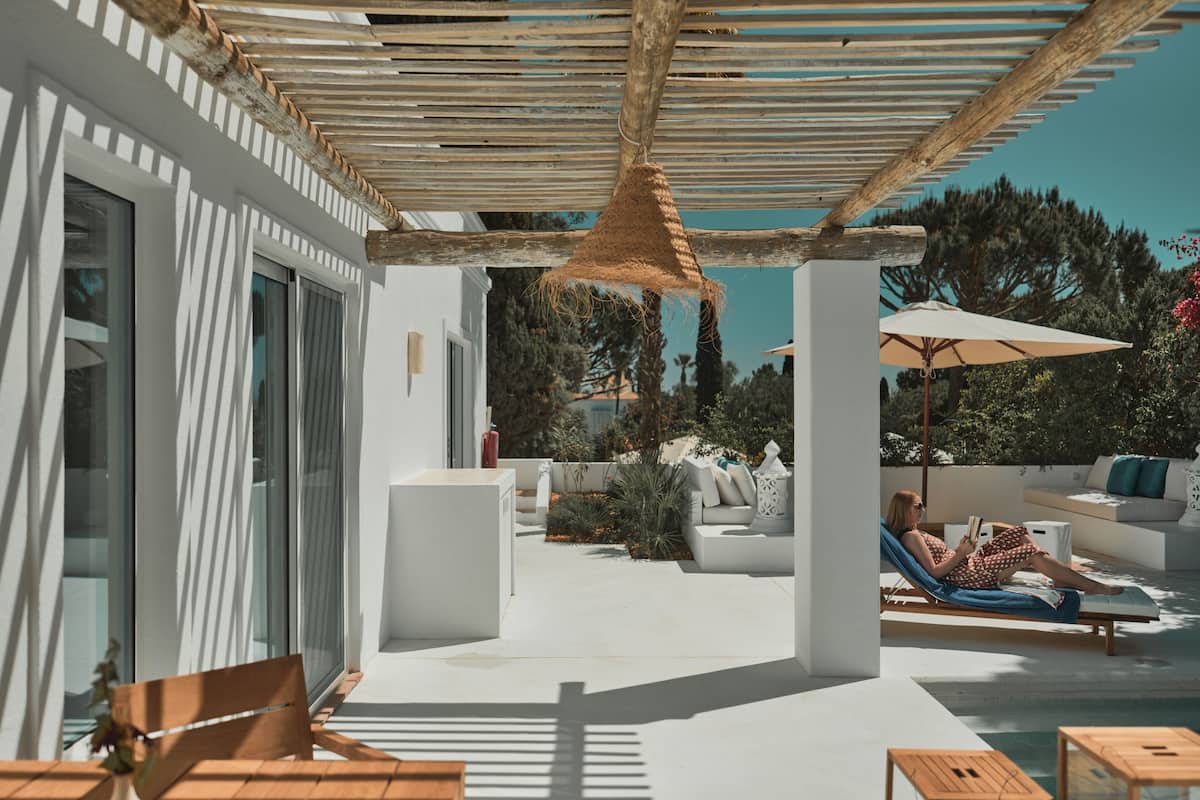 A modern outdoor lounge area with a pergola, seating, and a person reading a book.