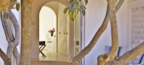 An interior hallway with a wooden floor, white walls, and a white door. There is a white chair and a small table with a vase of flowers.