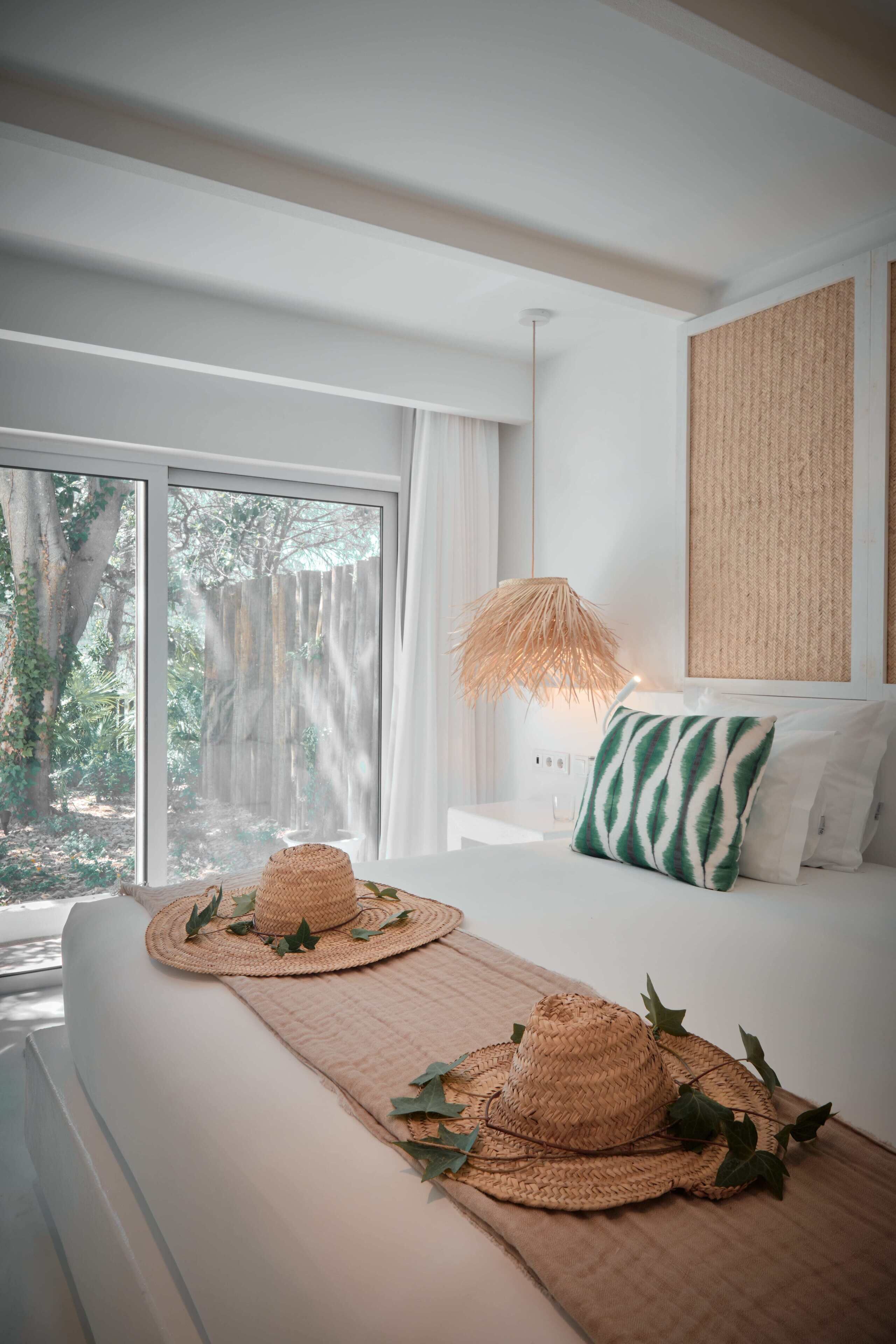 A bedroom with a large window, a bed with white bedding, a woven hat, and a woven mat.