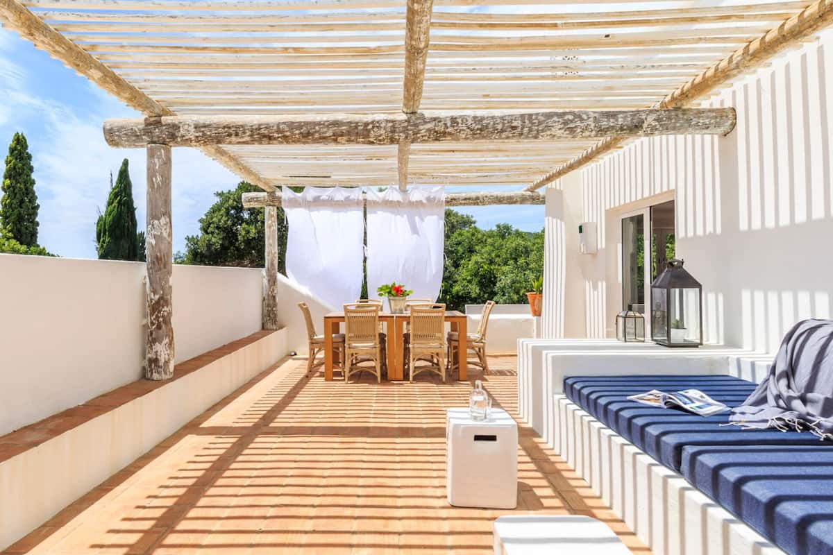 A covered outdoor area with a wooden table and chairs, a sofa, and a white speaker.