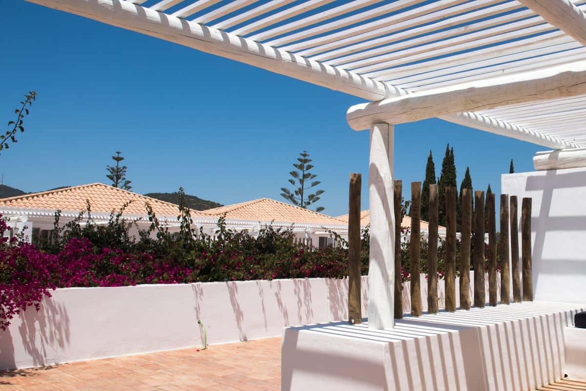 A white outdoor seating area with a pergola overhead, surrounded by a white wall and a row of trees.