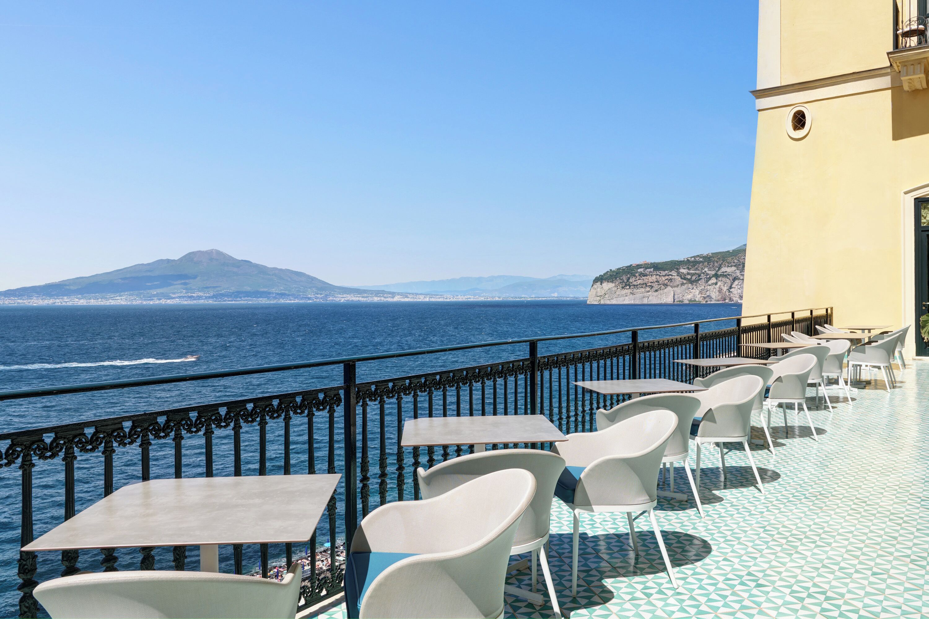 A terrace with tables and chairs overlooking the sea, featuring a mountain in the distance.