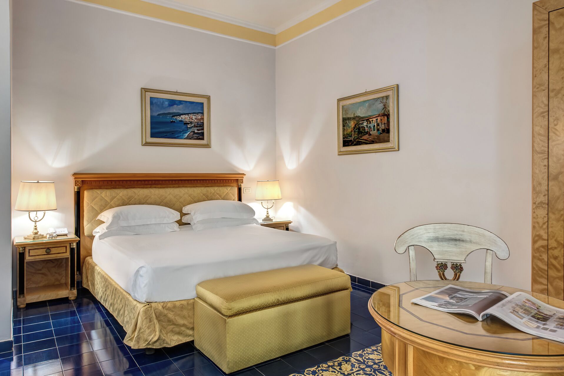 A hotel room with a large bed, two bedside lamps, a small table with a newspaper, and two framed pictures on the walls.