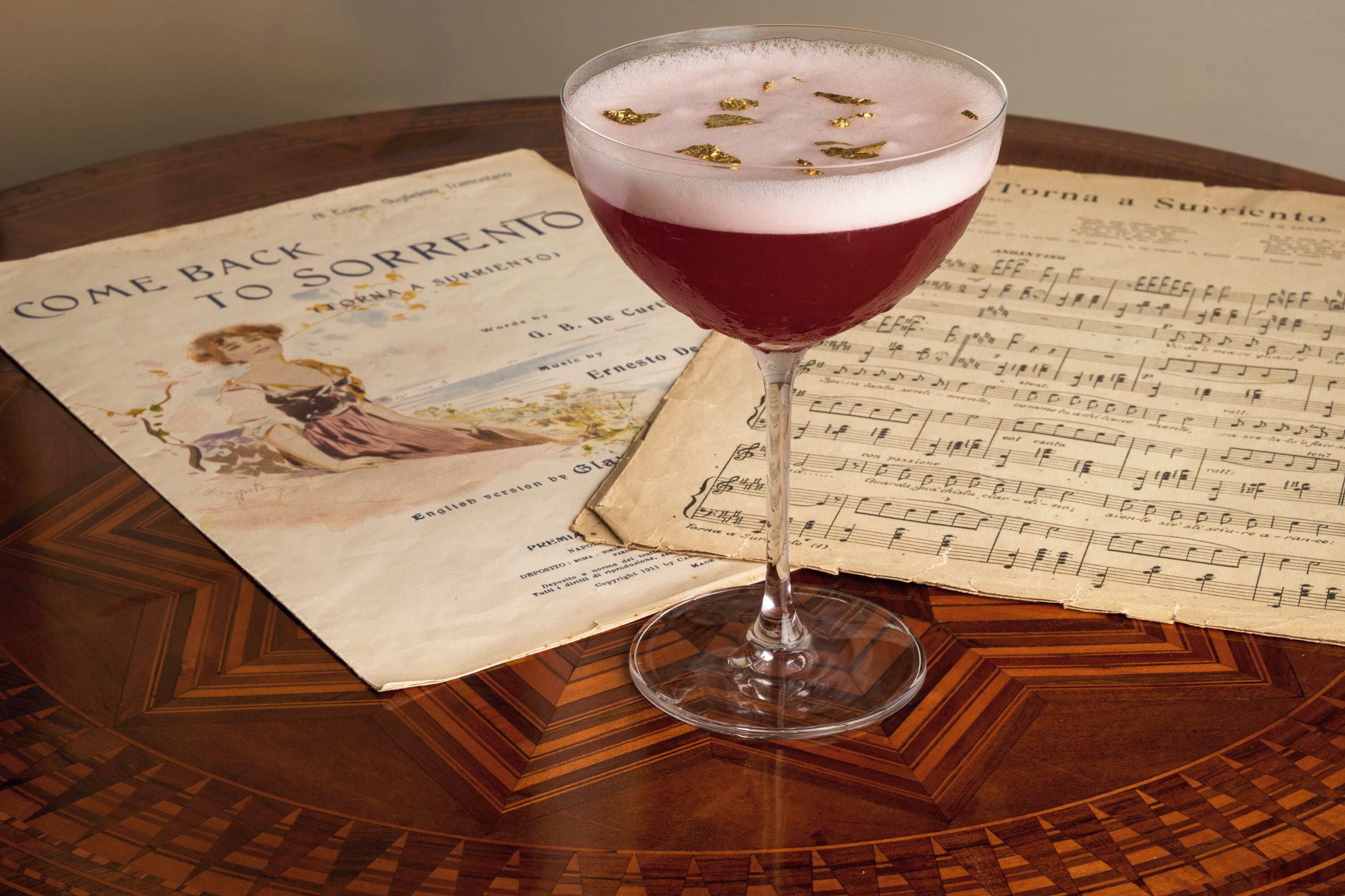 A glass of red beverage on a table with a sheet of music and a vintage poster.