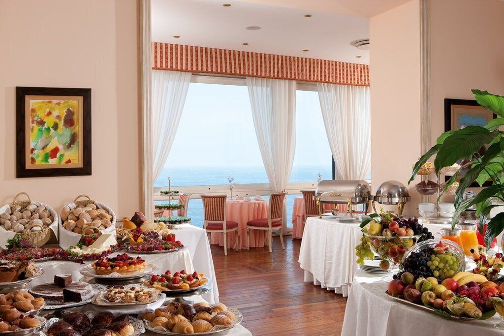 A breakfast buffet with various pastries, fruits, and beverages, set in a room with a view of the sea.