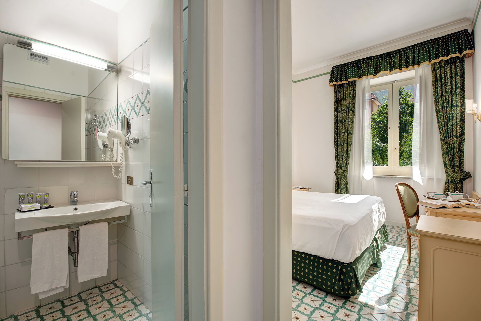 A hotel room with a bathroom, a bed with green bedding, a desk with a chair, and a window with curtains.