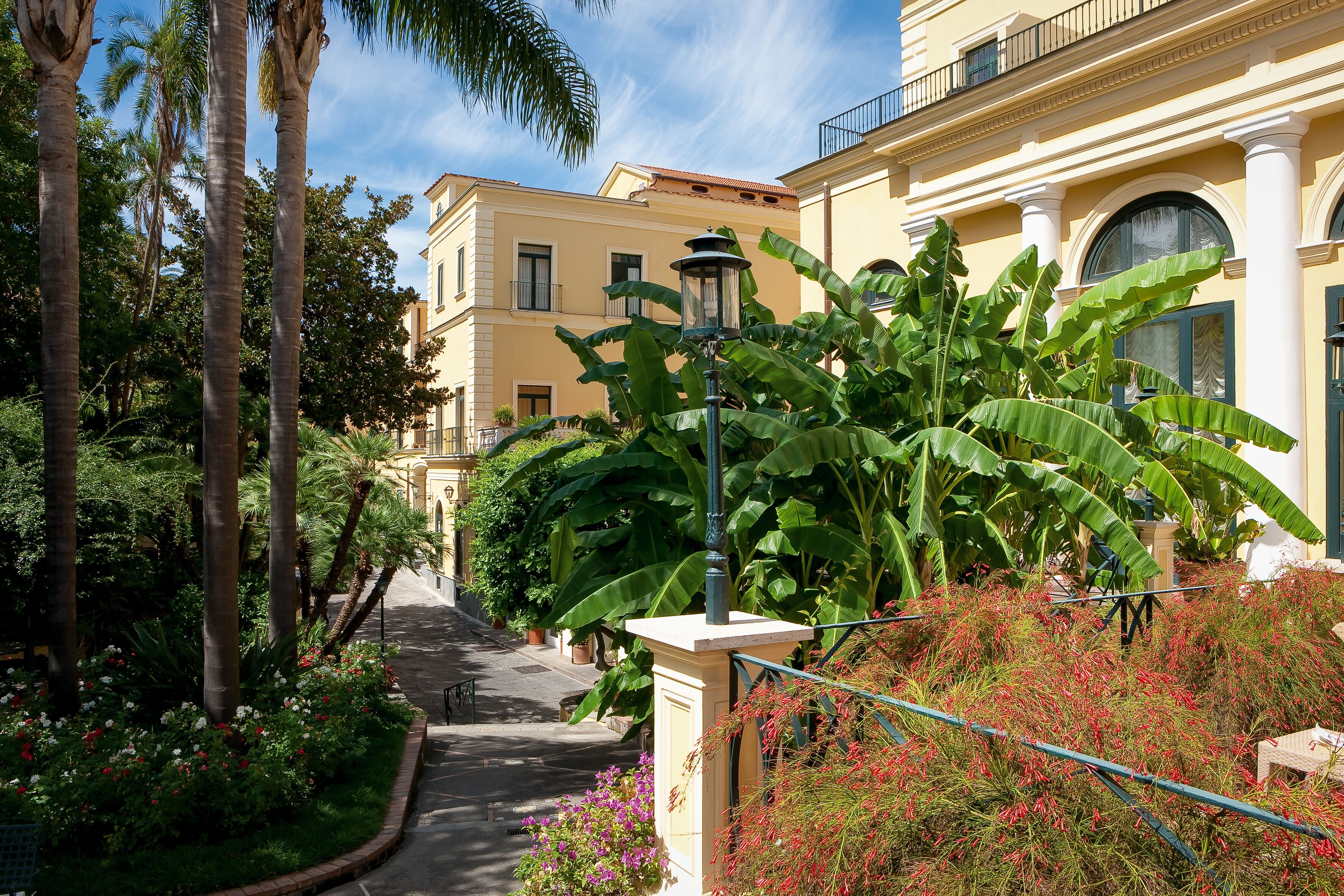 A narrow pathway flanked by lush greenery and a yellow building with columns and arched windows.