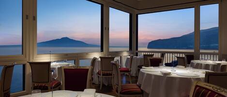 A restaurant with tables set for dining, large windows offering a view of the sea and mountains.