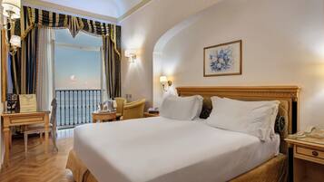 Habitación de hotel con una cama grande, un escritorio con teléfono, una silla y vista al mar a través de una ventana con cortinas.