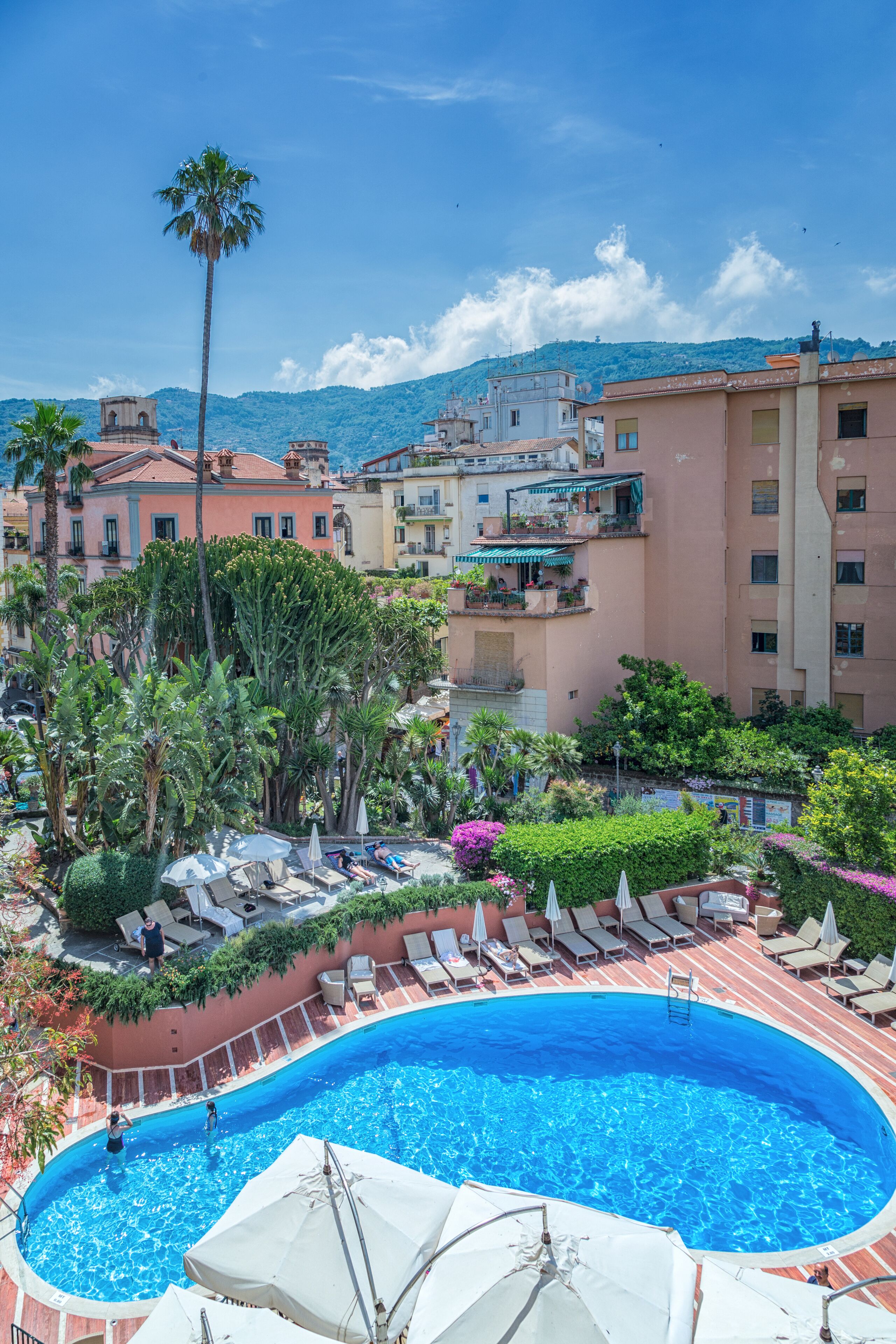 Una piscina sul tetto circondata da sedie a sdraio e ombrelloni, con edifici e una montagna sullo sfondo.