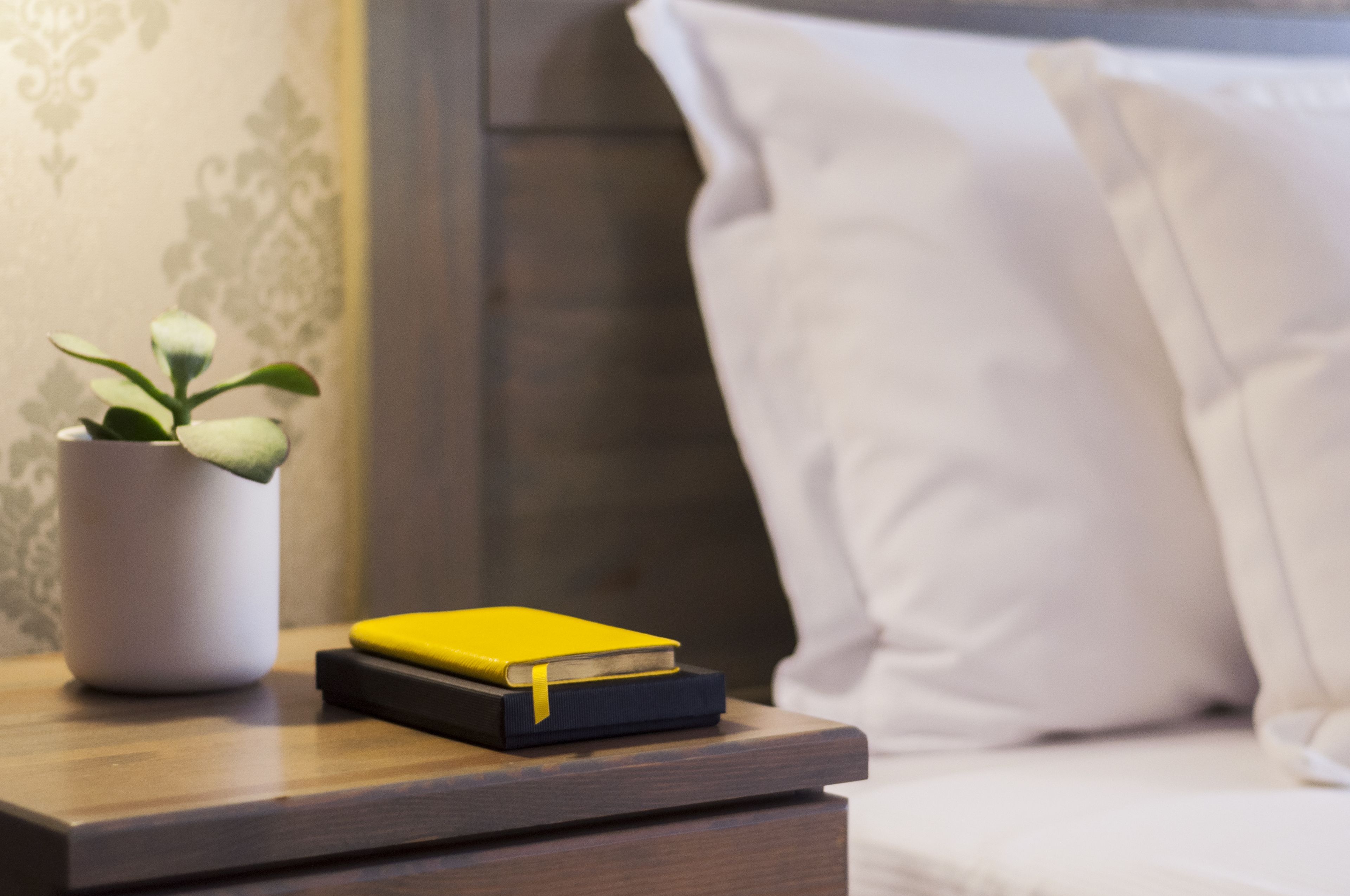 Une table de chevet avec une plante en pot et deux livres, un lit avec une literie blanche, et un mur à motifs en arrière-plan.