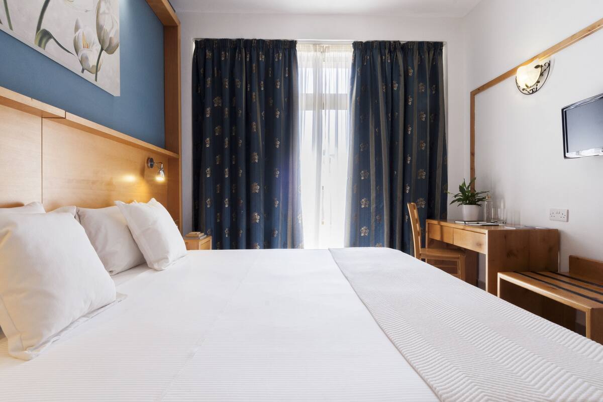 Hotellihuone, jossa on suuri sänky, työpöytä, tuoli, televisio ja ikkuna, jossa on verhot.