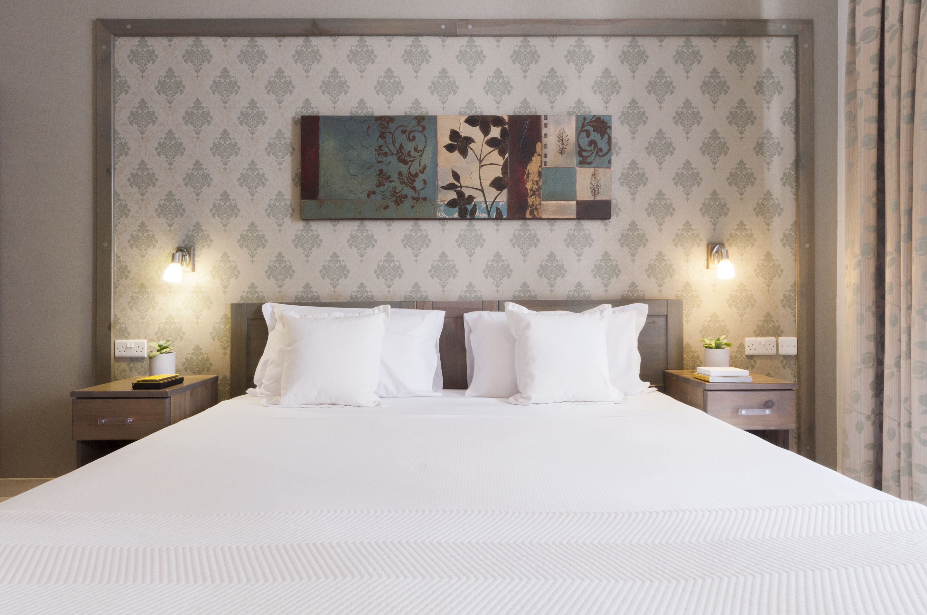 A hotel room with a large bed, two bedside tables, a wall-mounted artwork, and two wall-mounted lamps.