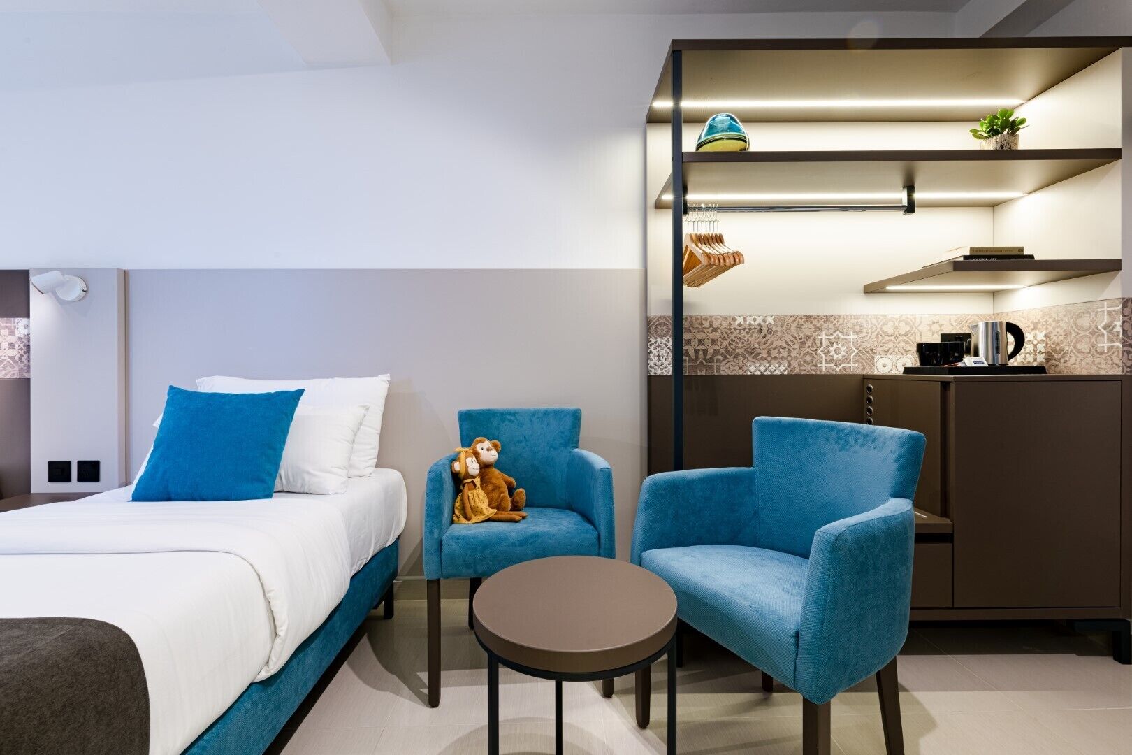 Une chambre d’hôtel moderne comprenant un lit, deux fauteuils bleus, une petite table ronde et un coin cuisine équipé d’une machine à café et agrémenté d’une plante.