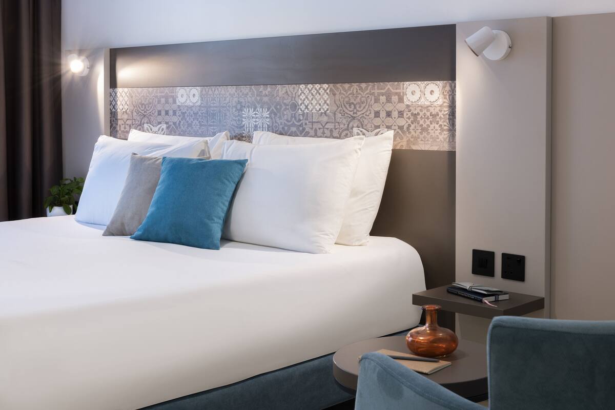 Une chambre d’hôtel moderne avec un lit soigneusement fait, une table de chevet ornée d’un vase et un luminaire fixé au mur.
