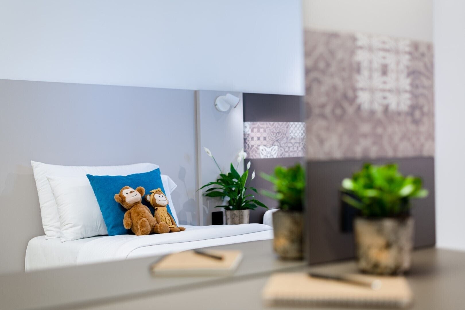 Una camera da letto moderna con un letto bianco, un cuscino blu, due morbidi peluche a forma di scimmia, un comodino con un libro e due piante in vaso.