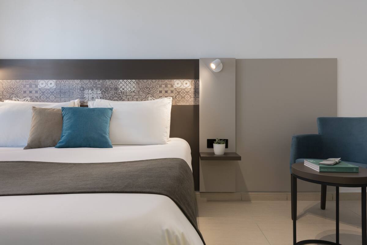 Une chambre d’hôtel avec un lit, une table de chevet ornée d’une plante, une chaise et une petite table sur laquelle se trouve un livre.