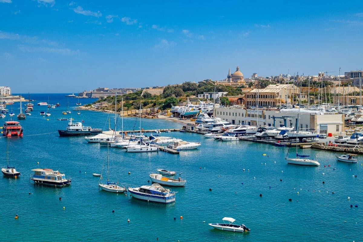 Un port de plaisance où de nombreux bateaux sont amarrés, un bâtiment imposant surmonté d’un dôme, et un ciel d’un bleu limpide.