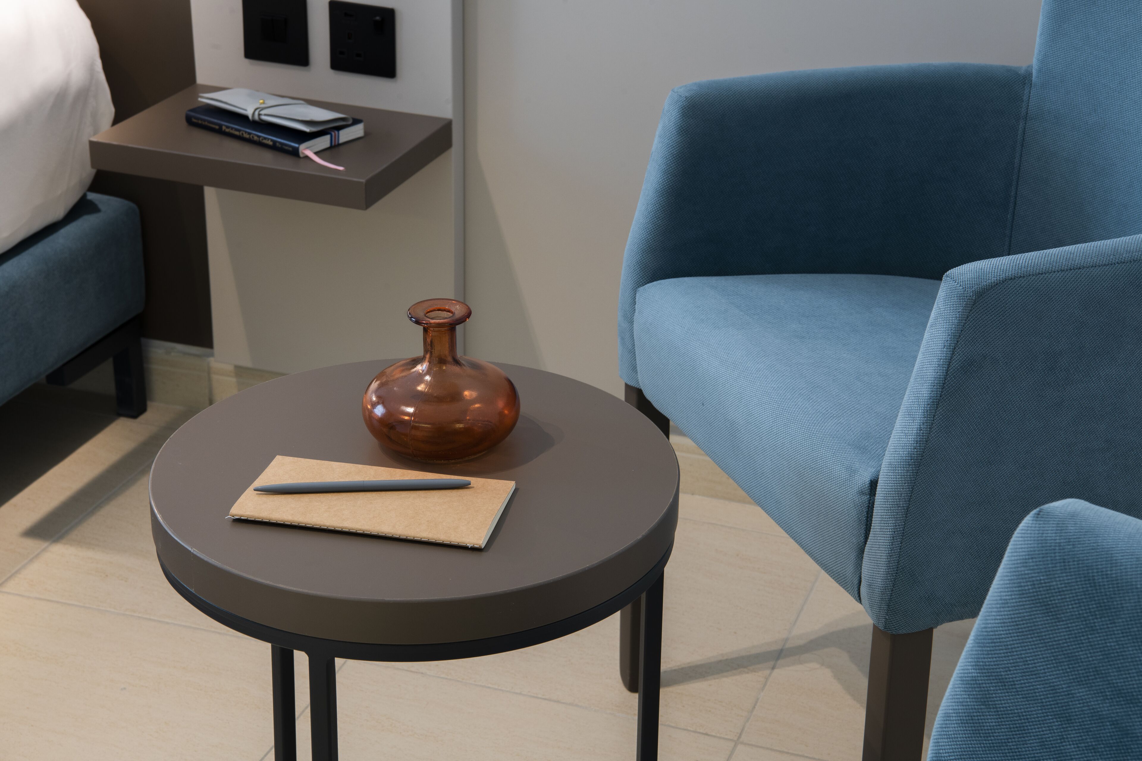 A modern room with a round table, a blue armchair, a small side table with a book and a pen, and a decorative vase.