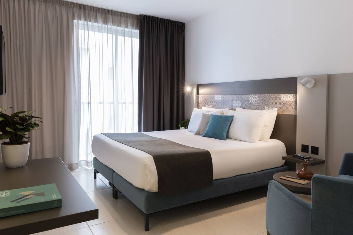 Une chambre d’hôtel moderne avec un grand lit, une table de chevet, une chaise, une plante en pot et une vue sur l’extérieur.