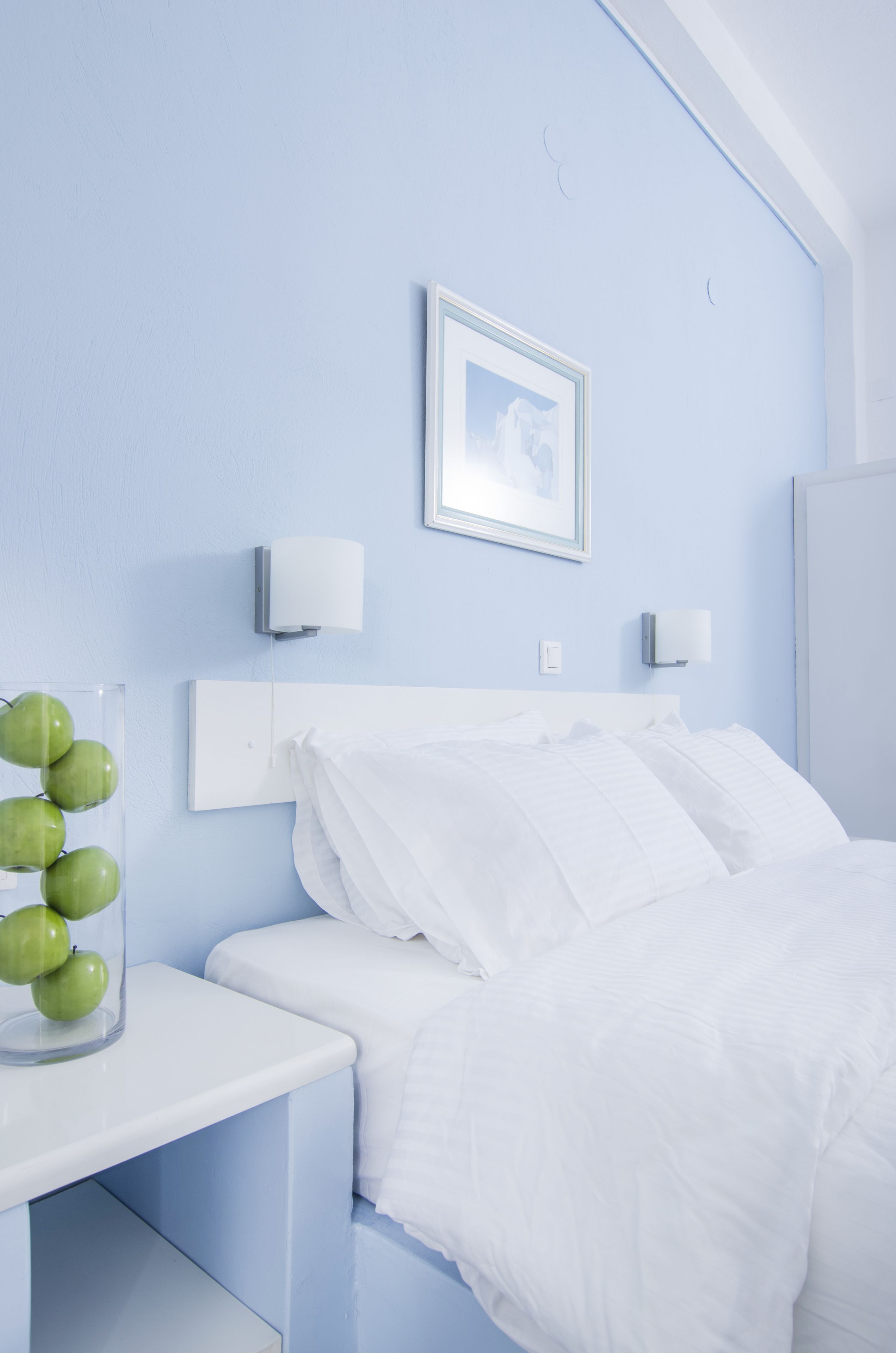 Un dormitorio con cama blanca, una mesita de noche con un florero de vidrio con manzanas verdes, y dos lámparas de pared.