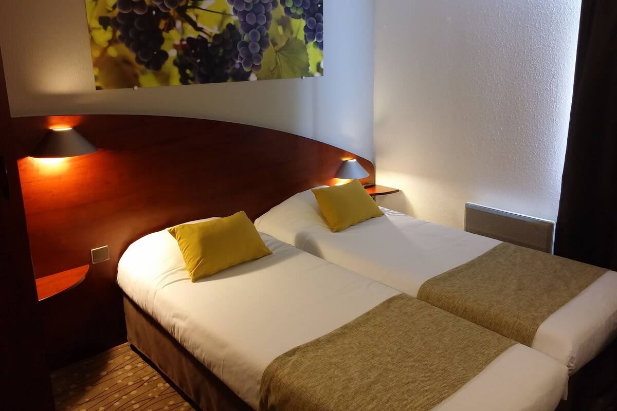 Une chambre d’hôtel avec deux lits, une grande peinture représentant des grappes de raisin et une tête de lit en bois.