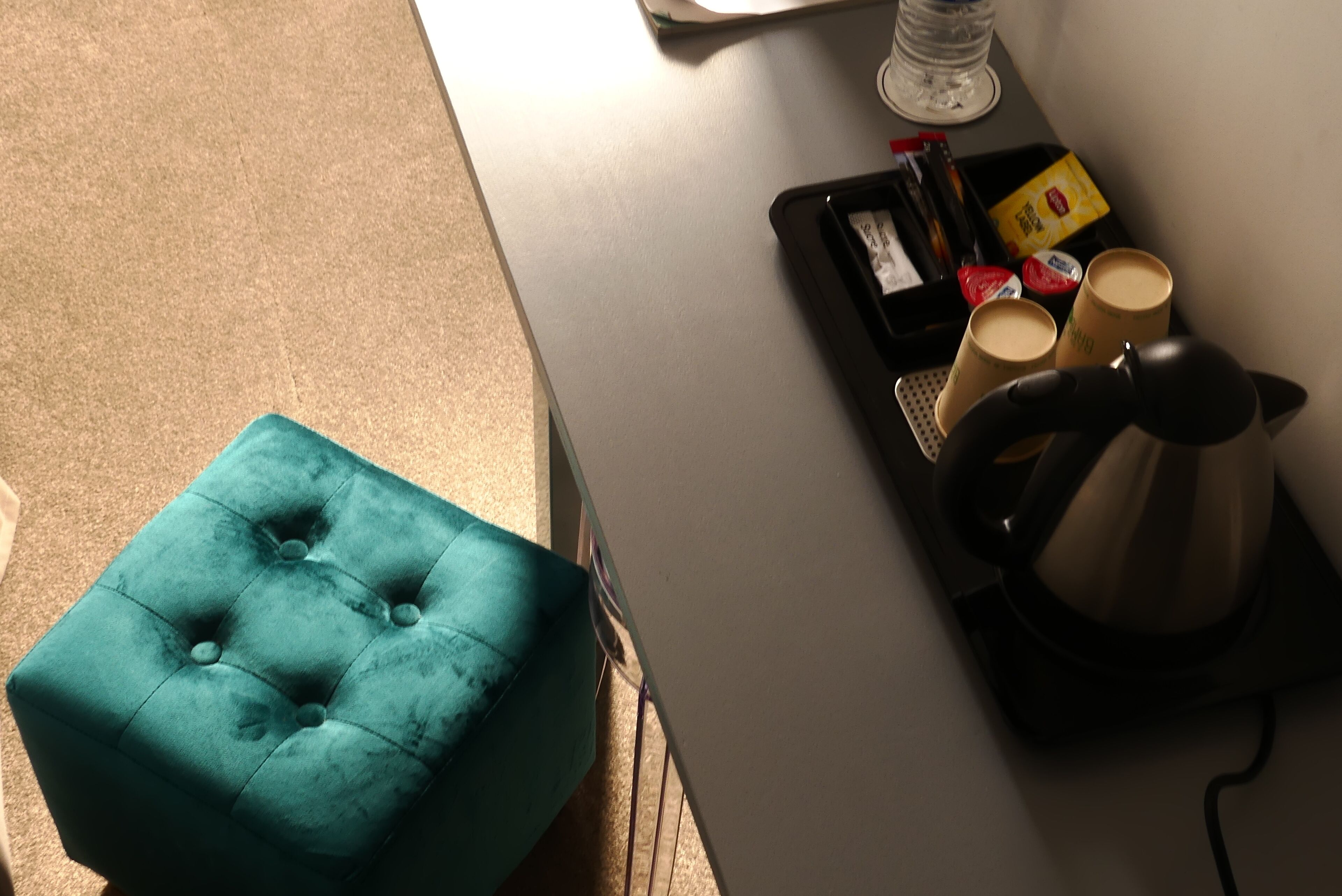 A teal ottoman, a black tray with a coffee maker, and two cups on a white table.