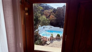 Vista desde una ventana a una piscina rodeada de árboles y un paisaje montañoso.