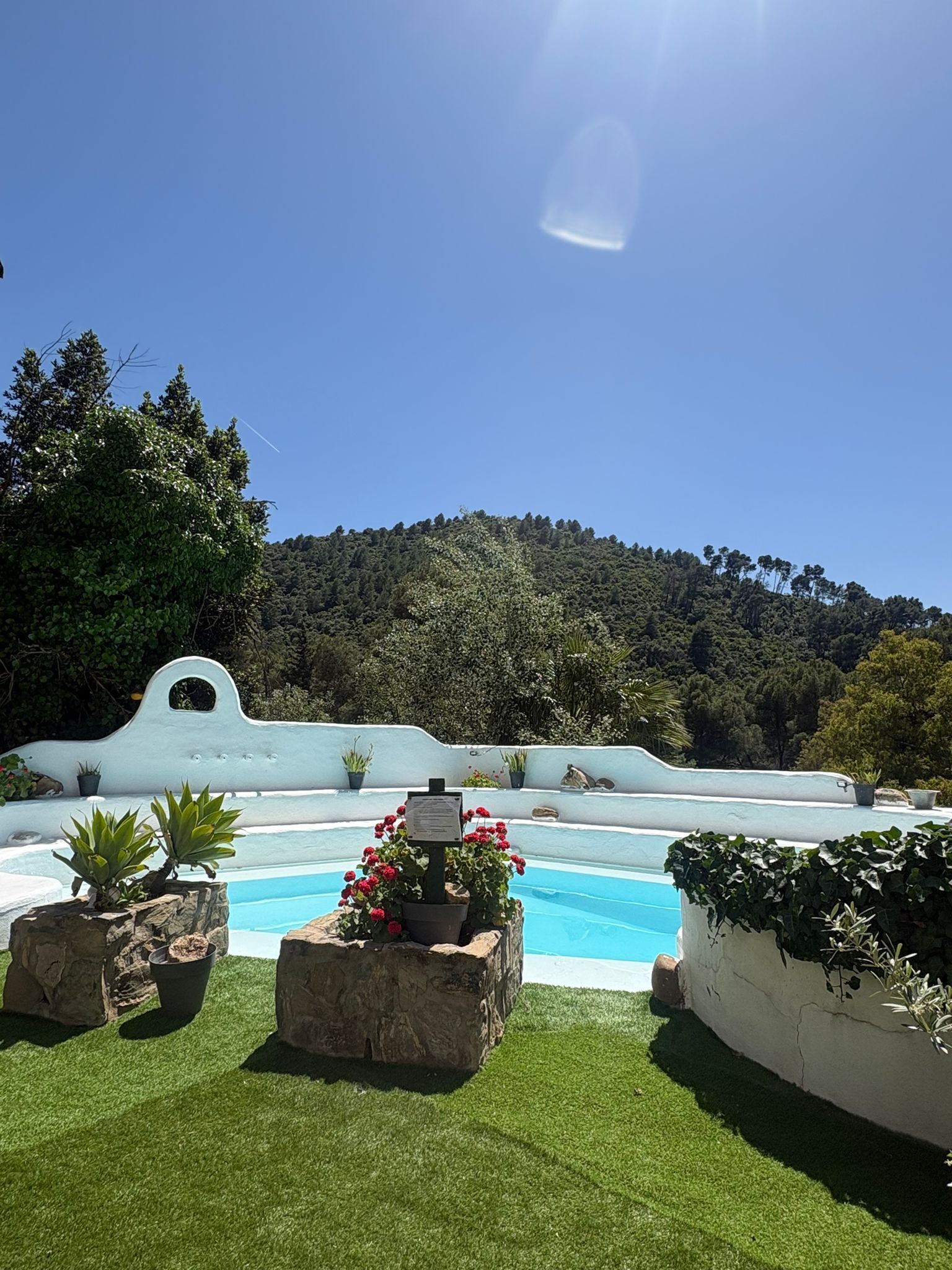 Una piscina amb una paret blanca, gespa verda i un turó al fons.