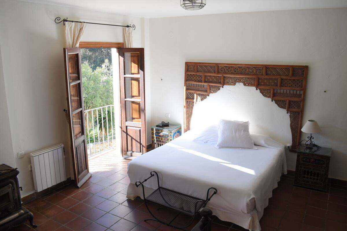 Ein Schlafzimmer mit einem großen Bett, einer geöffneten Balkontür, einem Heizkörper und einem traditionellen Holz-Kopfteil.