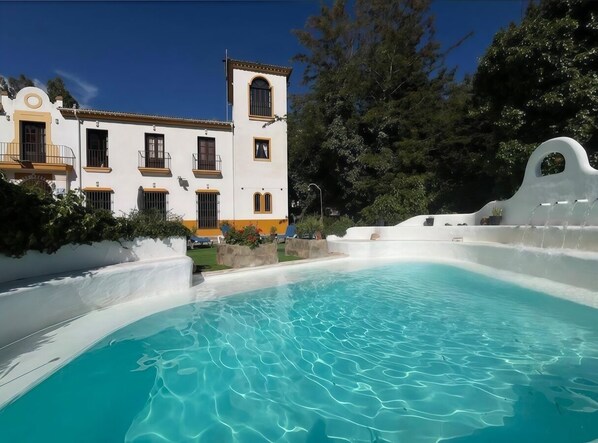 A pool with clear blue water, a white building with balconies, and a fountain.