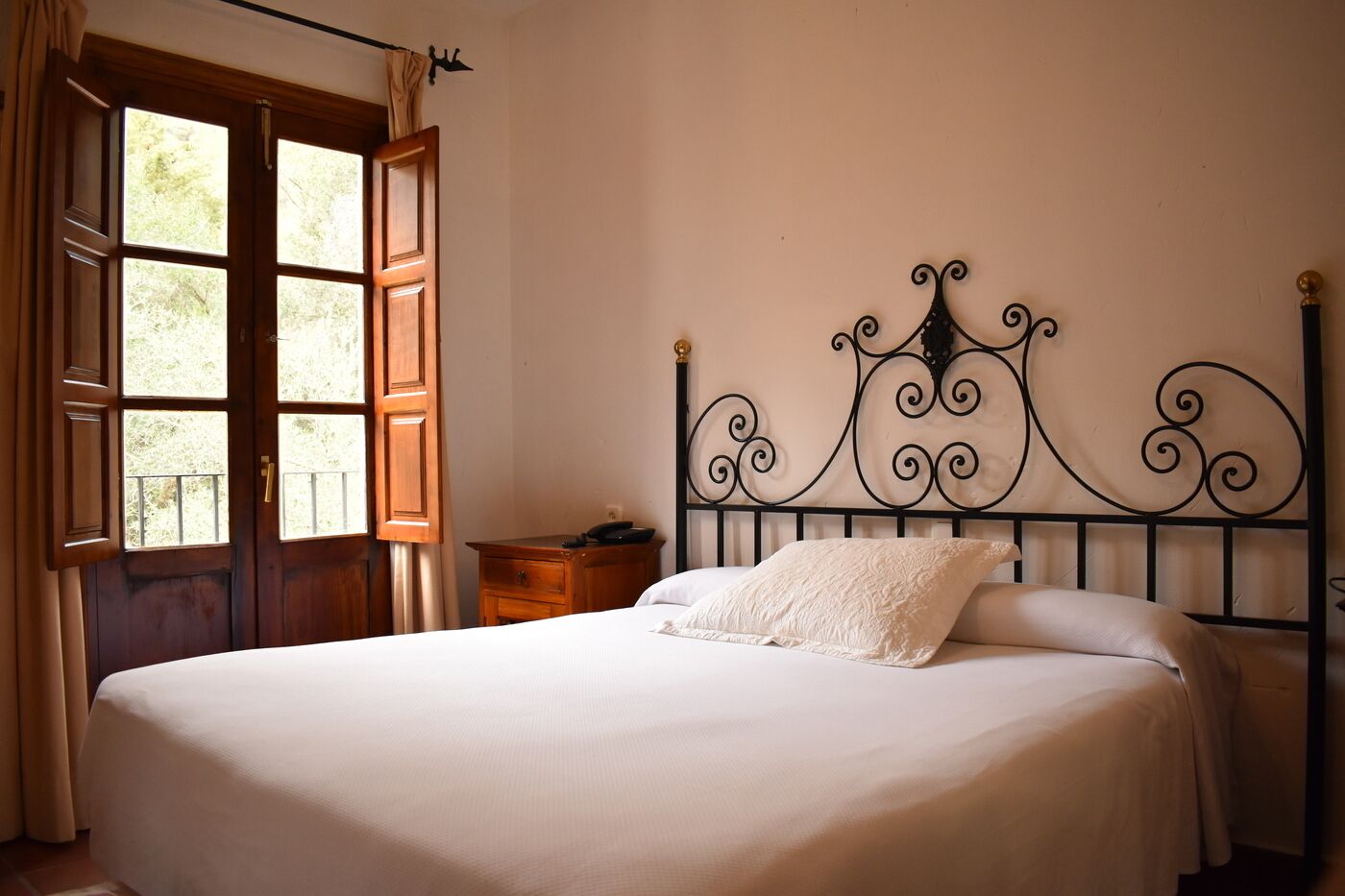 Een slaapkamer met een bed, een houten nachtkastje, een metalen hoofdbord en een raam met gordijnen.