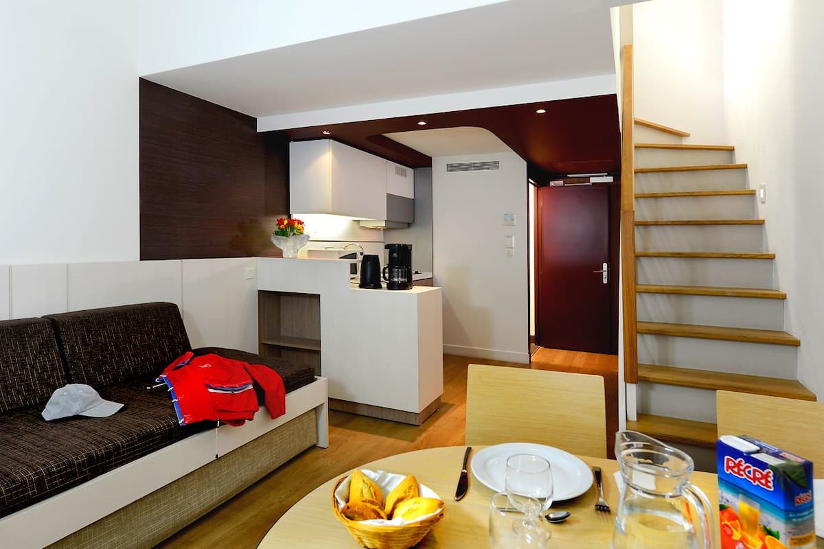 A modern hotel room with a sofa, a dining table with a basket of bread, a coffee maker, and a bottle of juice.