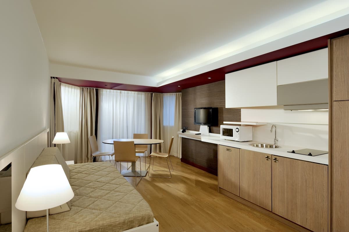 A hotel room with a bed, bedside lamp, a small table with chairs, a kitchenette with a microwave, and a TV.