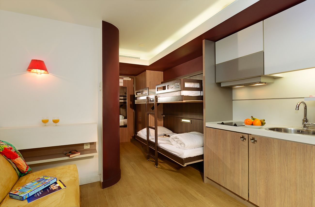 A compact hotel room with bunk beds, a kitchenette, a small seating area with a table, and a red wall lamp.