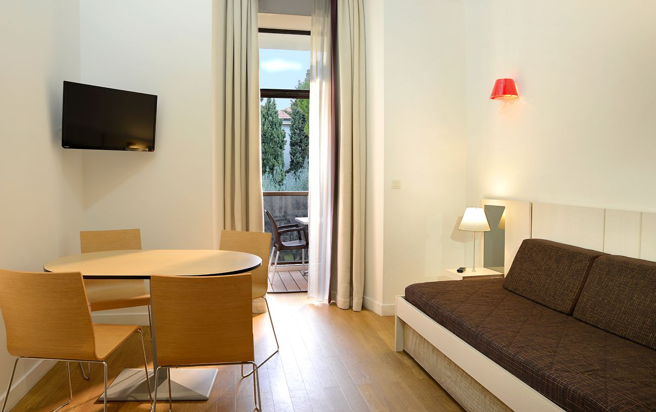 Una habitación de hotel moderna con comedor, sofá, televisor montado en la pared y vistas a través de una ventana.