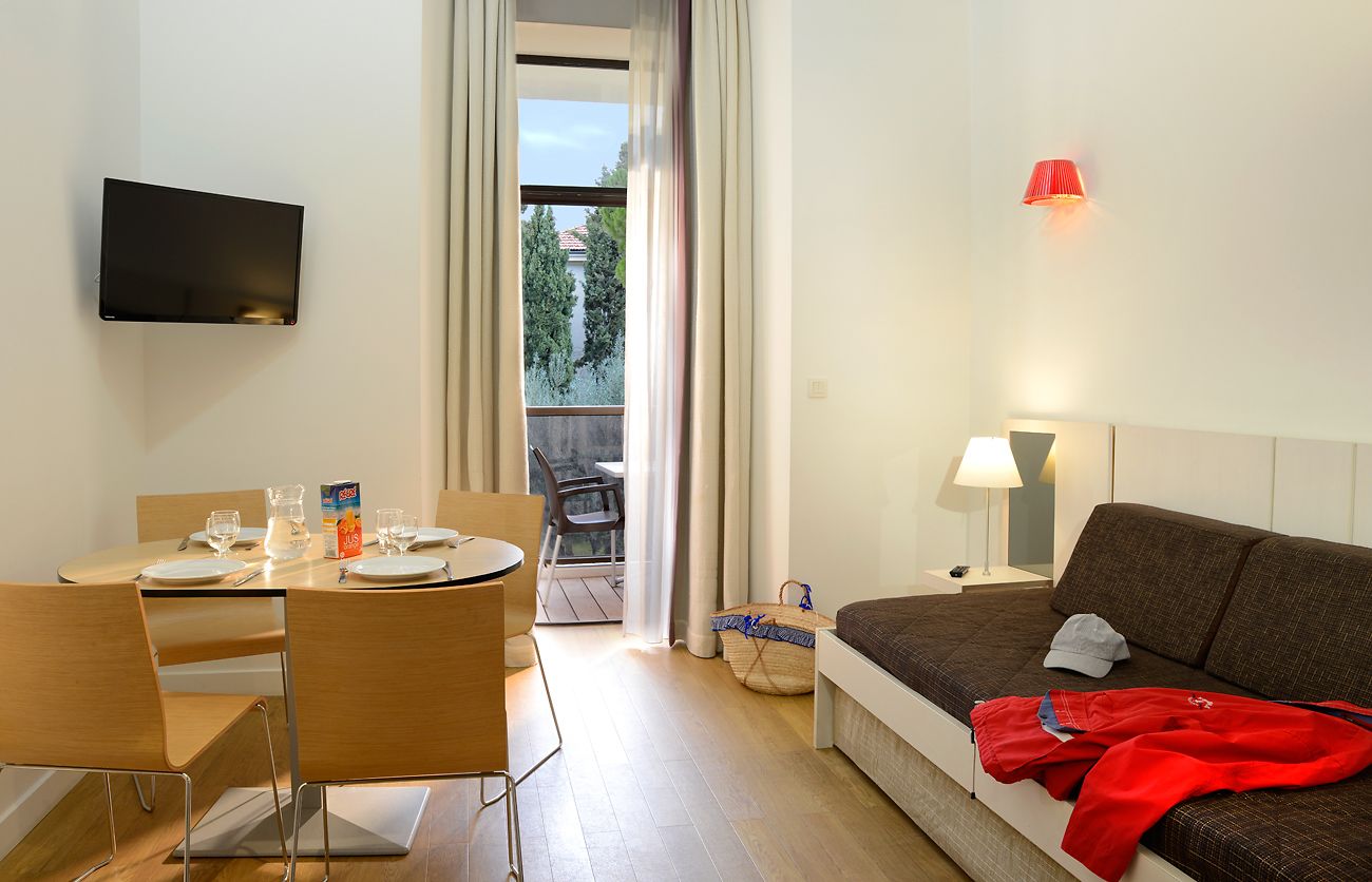 Una habitación de hotel moderna con una mesa de comedor preparada para dos personas, un sofá, un televisor montado en la pared y vistas a través de un amplio ventanal.