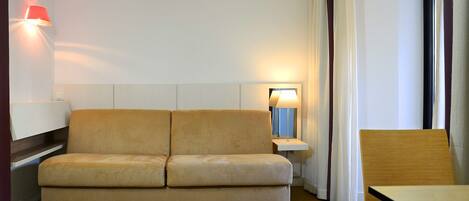 A modern hotel room with a beige sofa, a small side table with a lamp, a wooden chair, and a large window with curtains.