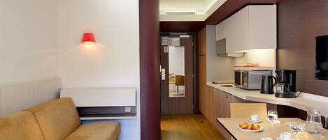 A modern hotel room with a sofa, a dining table with plates and glasses, a coffee maker, and a microwave.