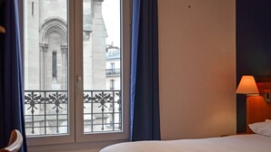A hotel room with a bed, a window with a view of a building, and a lamp on the nightstand. - Hôtel de la Perdrix Rouge (Paris)