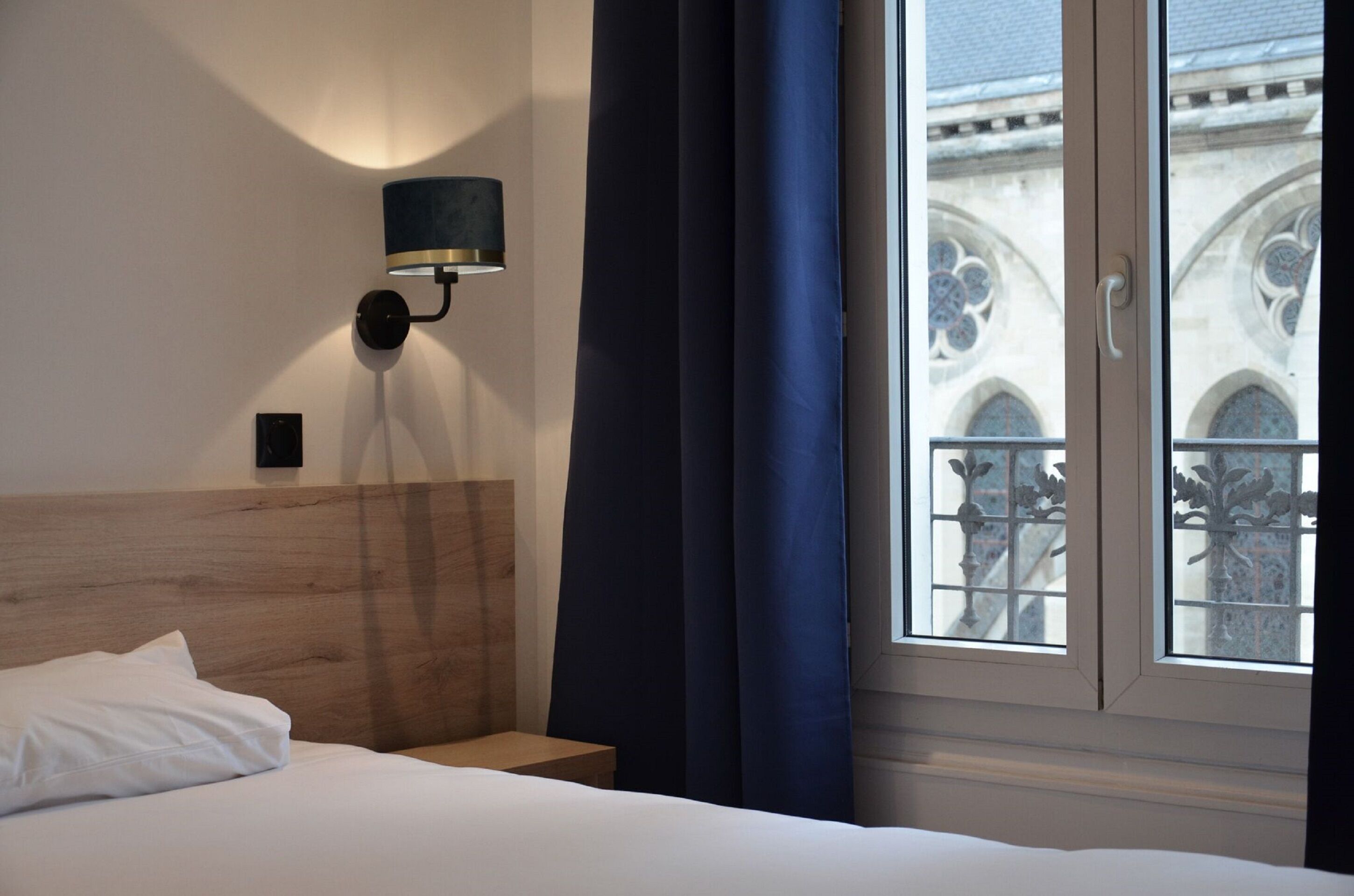 A hotel room with a wooden headboard, a bedside table, a lamp, and a window with a view of a building with ornate windows.