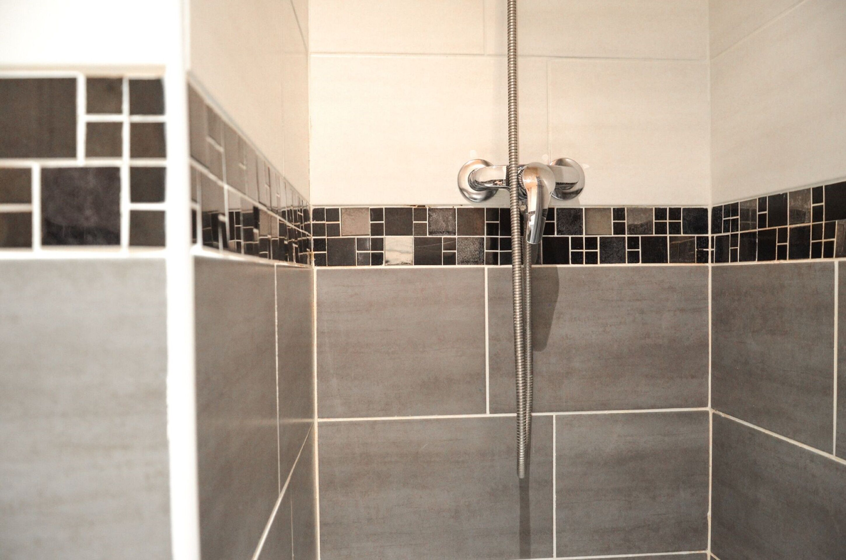 A shower area with a chrome showerhead and a flexible hose. The walls are tiled with a mix of large gray tiles and smaller black tiles.