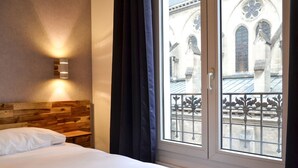 A hotel room with a wooden headboard, a bedside table, a bed with white linens, and a window with a view of a building with arched windows and a balcony with wrought iron railings. - Hôtel de la Perdrix Rouge (Paris)